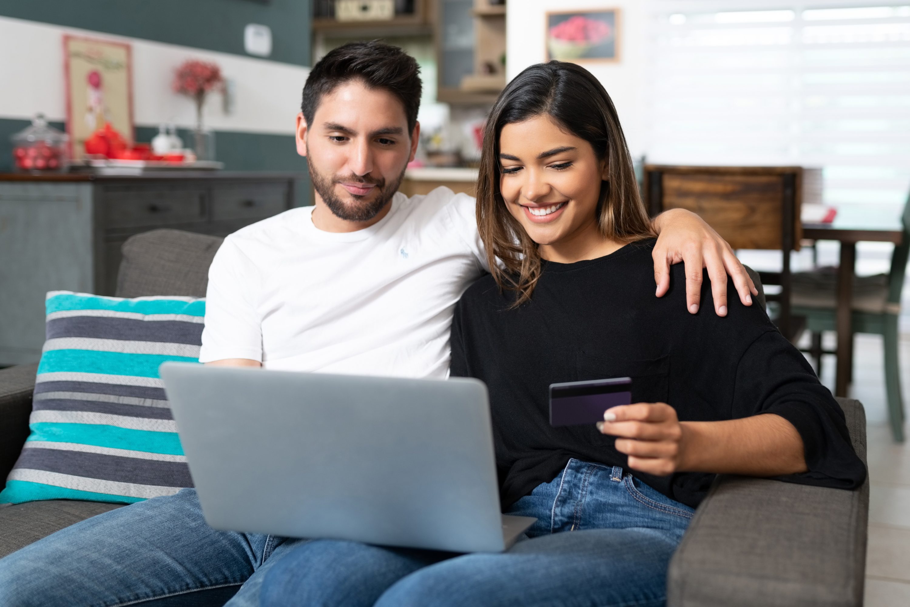 Happy couple shopping online through laptop while sitting on sofa in living room at home Happy couple shopping online through laptop while sitting on sofa in living room at home