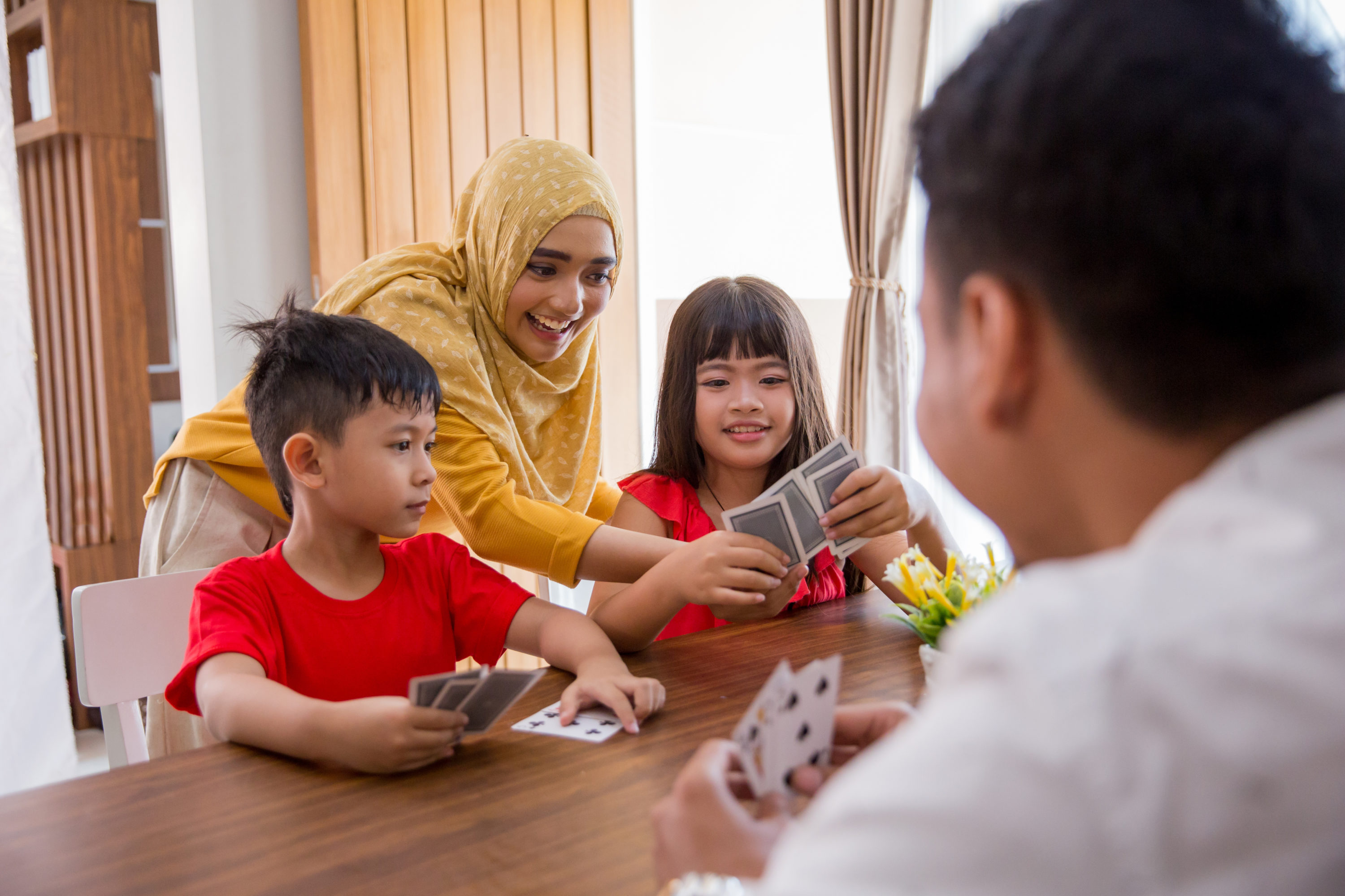 A family of four laughs together while playing cards at the kitchen table.