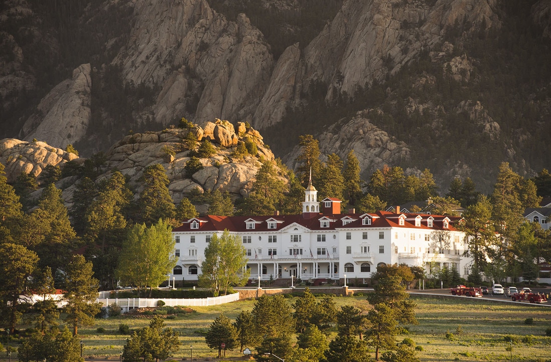 An aerial view The Stanley Hotel in Colorado. The hotel is set in front of mountains with trees in front.