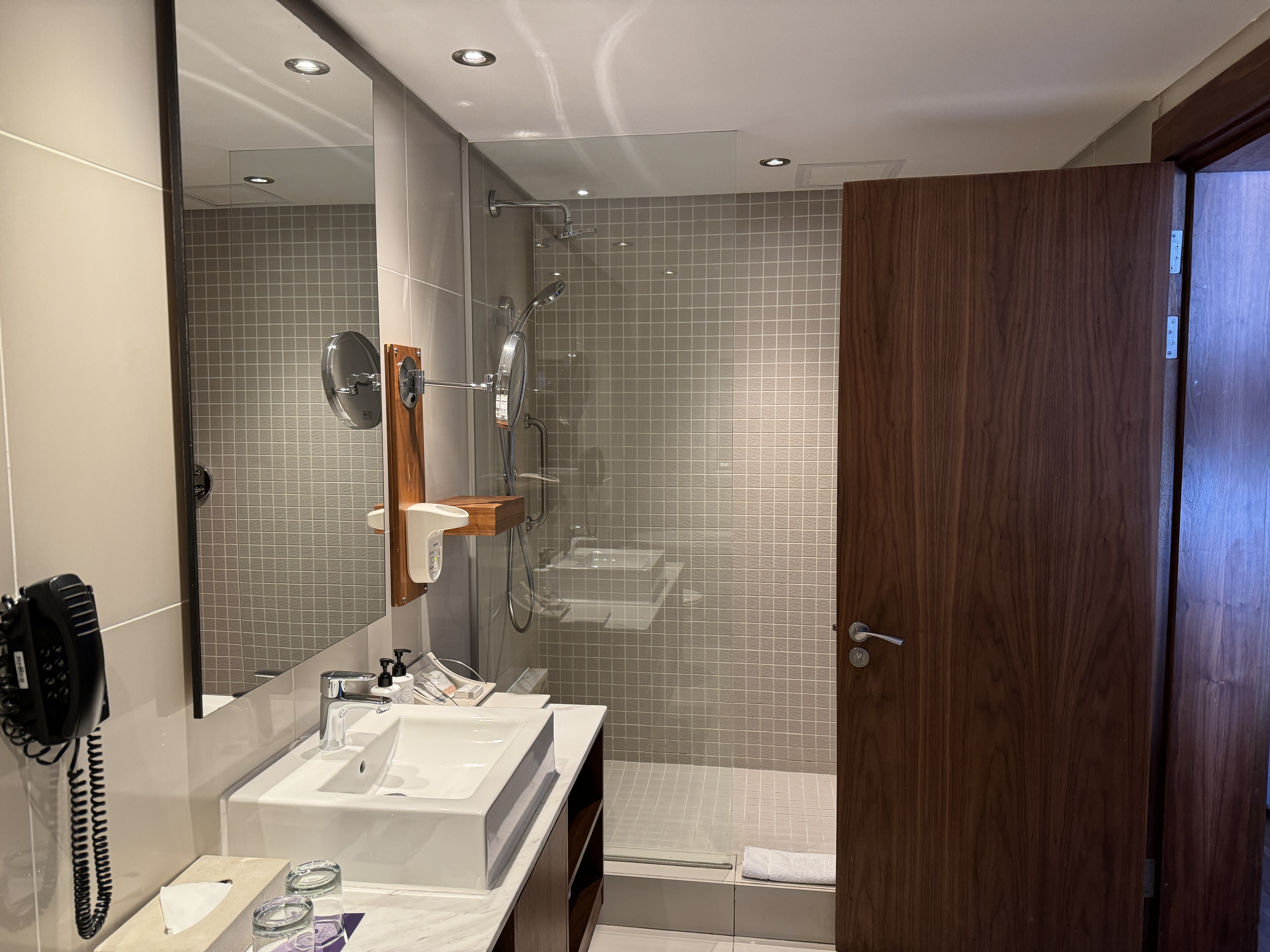 An image from inside of the bathroom with the shower in the background and singel sink in the foreground. The sink has 2 glasses, a tissue box, and full size body lotion and hand soap. An image from inside of the bathroom with the shower in the background and singel sink in the foreground. The sink has 2 glasses, a tissue box, and full size body lotion and hand soap.