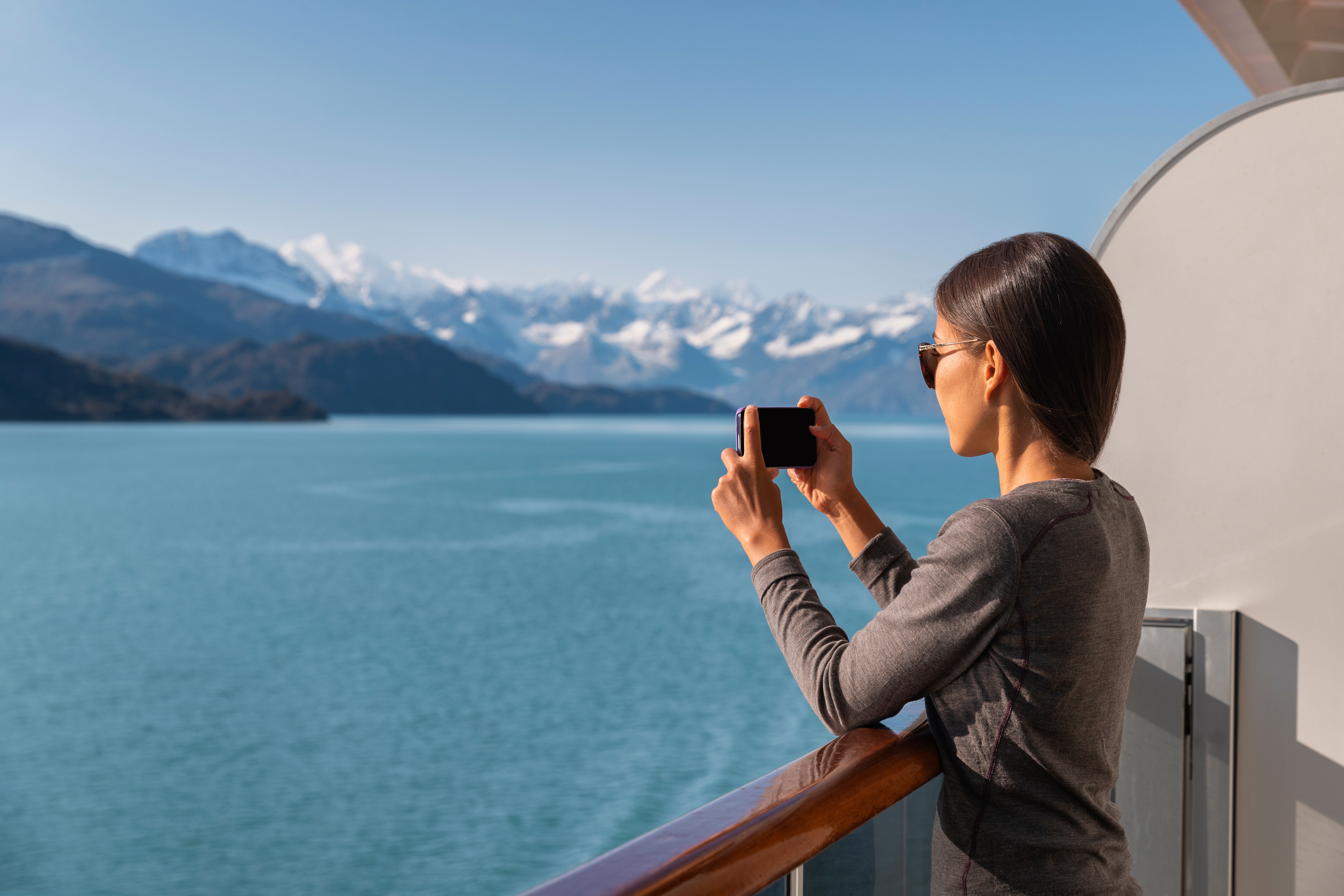 Alaska cruise ship passenger photographing amazing landscape entering Glacier Bay National Park, USA. Woman tourist taking photo picture using mobile cell smart phone on travel vacation Woman takes picture of glaciers in Alaska from cruise ship balcony